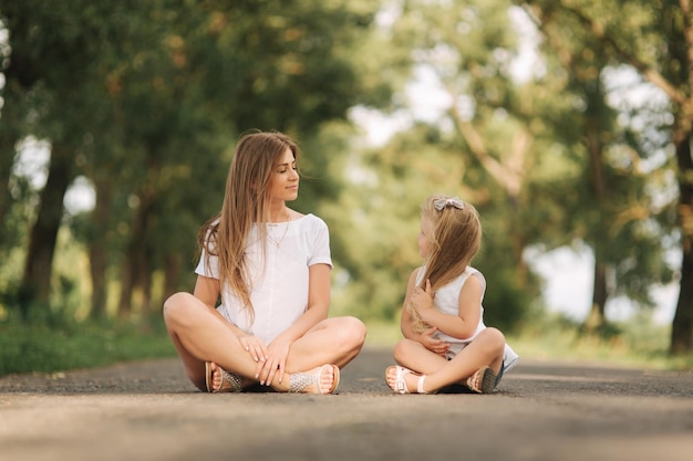 Atrakcyjna mama i córka o blond włosach siedzi na drodze w pobliżu dużej alei. Uśmiechają się i patrzą na natune