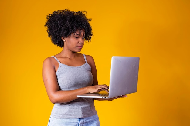 Atrakcyjna młoda afrykańska kobieta nosząca zwykłe ubrania stojąca na białym tle nad żółtym tłem przy użyciu laptopa