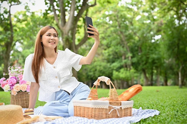 Atrakcyjna młoda Azjatka piknikuje w ogrodzie, używając smartfona do robienia selfie