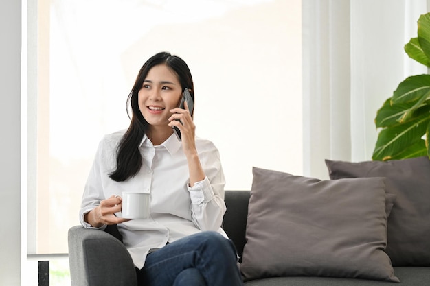 Atrakcyjna młoda Azjatka rozmawia przez telefon, relaksując się, popijając kawę na kanapie