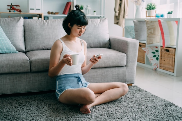 Atrakcyjna młoda azjatycka Japonka w domu na podłodze kryty. uśmiechający się ładna pani siedzi na kanapie opierając się dywan z inteligentny telefon w ręce podczas picia filiżankę herbaty. kobieta w skrócie w nowoczesnym przytulnym domu