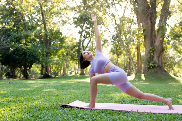 Atrakcyjna młoda Azjatycka kobieta praktykuje ćwiczenia jogi w parku, stojąc jedną nogę na macie do jogi, pokazując równowagę postawy Koncepcja życia i aktywności dobrego samopoczucia