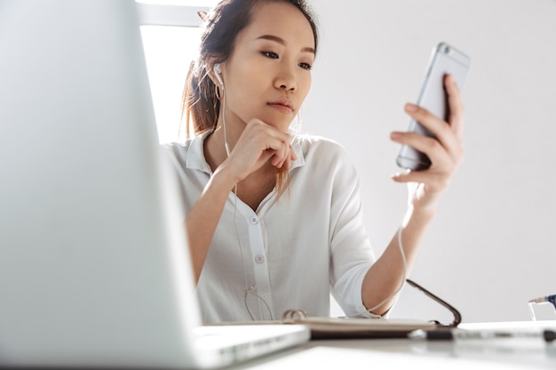 Atrakcyjna młoda bizneswoman azjatyckiego siedzi w biurze z komputera przenośnego trzymając telefon komórkowy, na sobie słuchawki