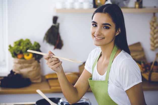 Zdjęcie atrakcyjna młoda ciemnowłosa kobieta degustująca gotowy gorący posiłek z drewnianą łyżką, stojąc i uśmiechając się w pobliżu kuchennego pieca koncepcje gotowania i prowadzenia gospodarstwa domowego