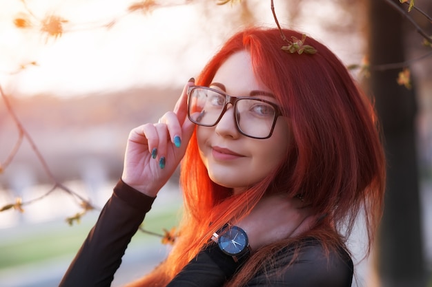Atrakcyjna młoda dziewczyna z rudymi włosami, w okularach, pozuje w pobliżu drzewa o wschodzie słońca