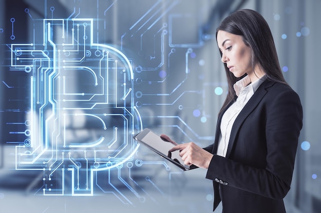 Atrakcyjna młoda europejska kobieta za pomocą tabletu z kreatywnym świecącym hologramem obwodu bitcoin na tle rozmazanego pokoju biurowego Kryptowaluta blockchain i koncepcja finansów Podwójna ekspozycja