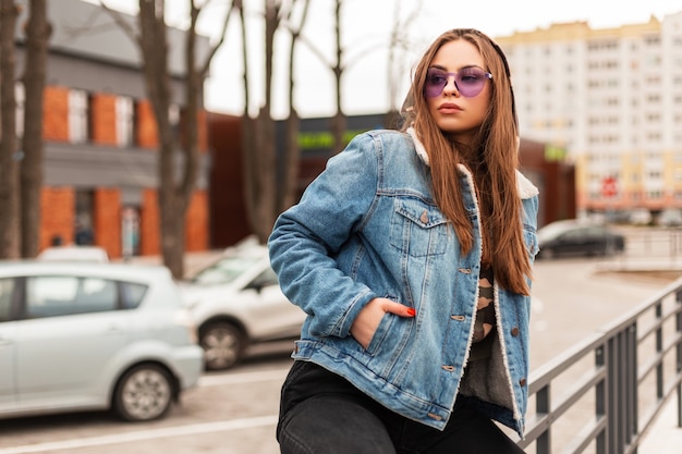 Atrakcyjna Młoda Hipster Kobieta W Stylowych Fioletowych Okularach W Wojskowym Kapturze W Modnej Niebieskiej Kurtce Dżinsowej Relaksuje Się Na Ulicy W Wiosenny Dzień. Piękna Dziewczyna Miejska Cieszy Się Na Wakacje W Mieście.