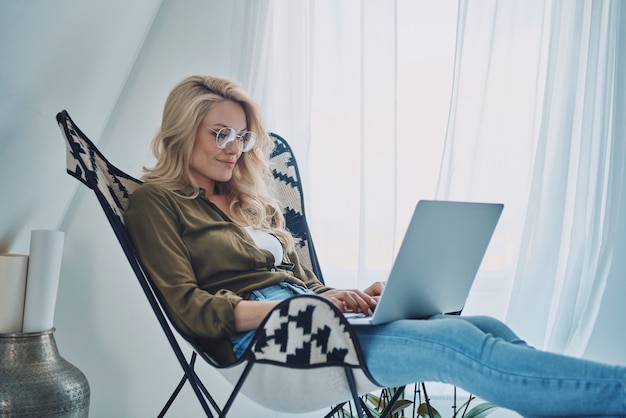 Atrakcyjna młoda kobieta korzysta z laptopa siedząc w wygodnym fotelu w domu