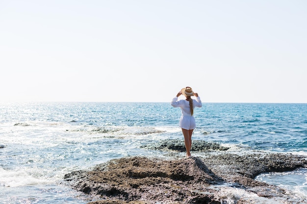 Atrakcyjna młoda kobieta o długich blond włosach w białym letnim garniturze stoi nad brzegiem morza i cieszy się widokiem na ocean.