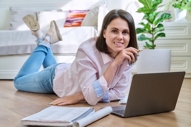 Atrakcyjna młoda kobieta patrząca na kamerę leżącą na podłodze w domu z laptopem
