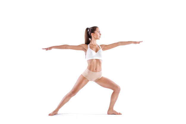 Atrakcyjna młoda kobieta robi ćwiczenia jogi Virabhadrasana, na białym tle. Ćwiczenia kontrolujące równowagę, studyjne pełnej długości.