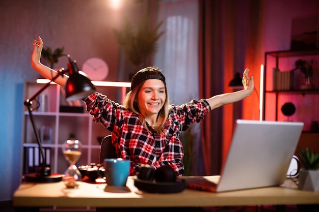 Atrakcyjna młoda kobieta siedzi przy biurku z podnoszeniem rąk i patrząc na ekran laptopa