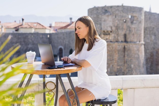 Atrakcyjna młoda kobieta siedzi przy stoliku przed kawiarnią i pracuje na laptopie