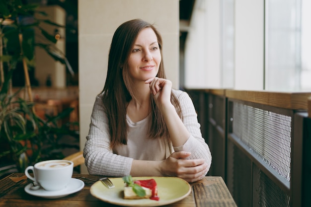 Atrakcyjna Młoda Kobieta Siedzi Sama W Pobliżu Duże Okno W Kawiarni Przy Stole Z Filiżanką Cappuccino, Ciasto, Relaks W Restauracji W Czasie Wolnym. Młoda Kobieta Po Odpoczynku W Kawiarni. Koncepcja Stylu życia.