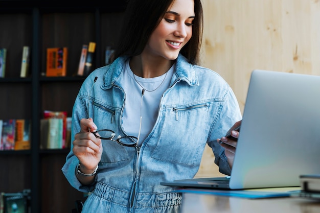 Atrakcyjna młoda kobieta stoi przy oknie, trzyma w ręku smartfon, okulary do widzenia i pracuje na laptopie.