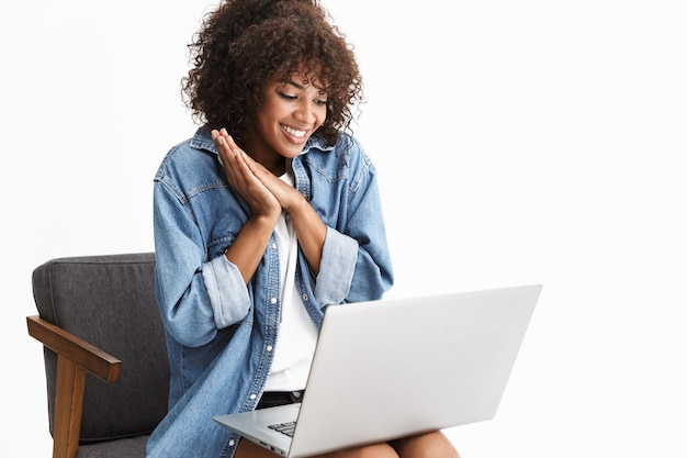 Atrakcyjna młoda kobieta ubrana w dżins, siedząca na krześle na białym tle nad białą ścianą, pracująca na laptopie, rozmawiająca