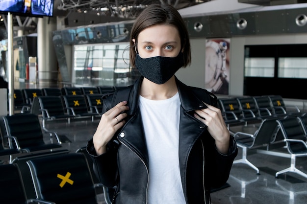 Atrakcyjna Młoda Kobieta W Czarnej Masce Ochronnej Z Plecakiem Na Terminalu Lotniska Międzynarodowego. Podróżowanie Podczas Pandemii Koronawirusa.