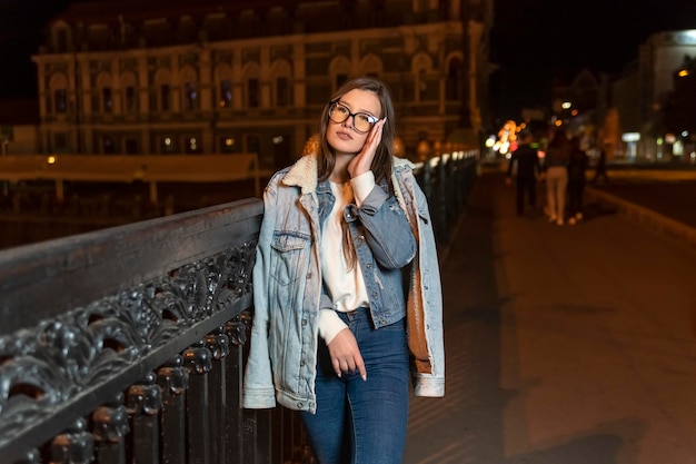 Atrakcyjna młoda kobieta w okularach i dżinsowym stylu na tle miasta wieczorem Stylowa dziewczyna spaceruje nocą po ulicy miasta