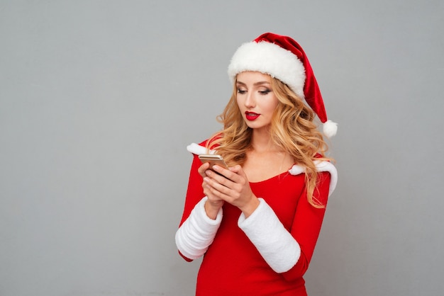 Atrakcyjna Młoda Kobieta W Stroju świętego Mikołaja Stojąca I Używająca Telefonu Komórkowego Na Szarej Powierzchni