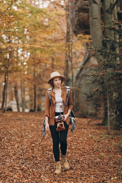 Atrakcyjna młoda kobieta w stylowym kapeluszu i torbie podróżnej na ramionach, rozglądająca się po uroczym jesiennym lesie
