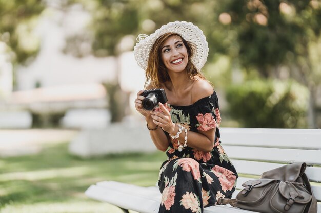 Atrakcyjna młoda kobieta z aparatem cyfrowym, ciesząc się letnimi wakacjami, siedząc na ławce w parku.