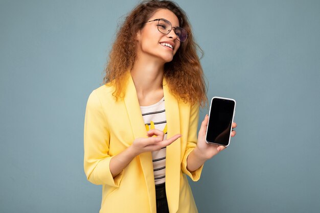 Atrakcyjna młoda kobieta z kręconymi ciemnymi blond włosami, ubrana w żółtą kurtkę i okulary optyczne na białym tle, trzymając i pokazując telefon komórkowy z pustym miejscem na wycięcie