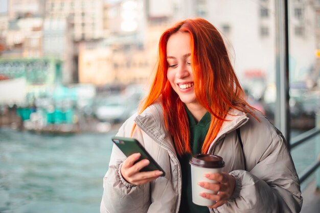 Zdjęcie atrakcyjna młoda rudowłosa kobieta wpisuje wiadomość przez telefon komórkowy na promie w stambule i się uśmiecha