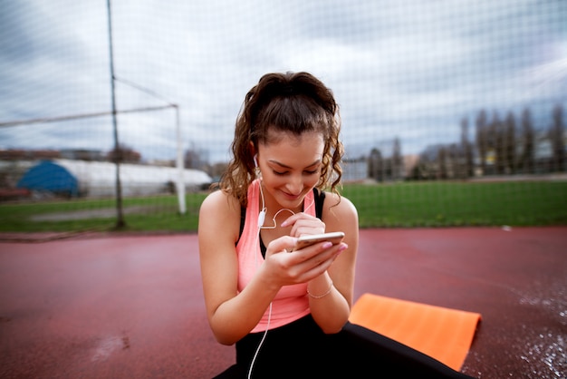 Atrakcyjna młoda sportive kobieta sprawdza telefon podczas gdy siedzący na pomarańczowej macie.