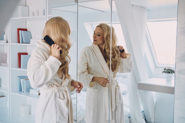 Atrakcyjna młoda uśmiechnięta kobieta w szlafroku, czesząca włosy, stojąc przed lustrem w domu