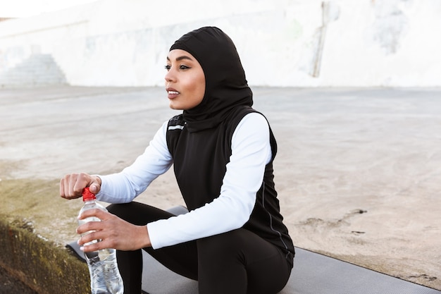 Atrakcyjna Muzułmańska Sportsmenka Nosząca Hidżab Na Zewnątrz, Siedząca Na Macie Fitness, Pijąca Wodę