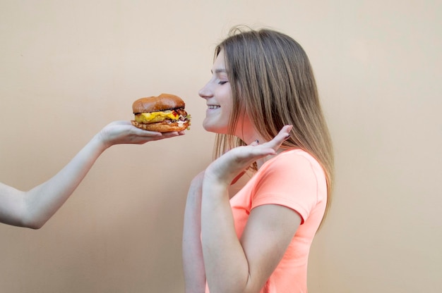 Atrakcyjna nastolatka stoi przy pomarańczowej ścianie w pomarańczowej koszulce i wącha dużego burgera