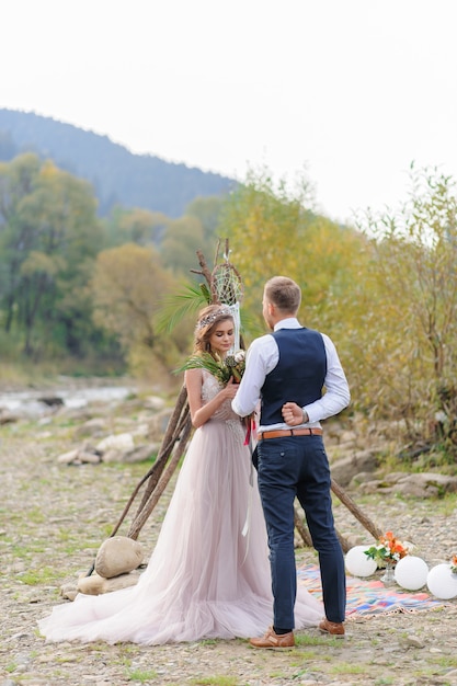 Atrakcyjna nowożeńcy, szczęśliwa i radosna chwila. Mężczyzna daje kwiaty ukochanemu. Ślub Cermonia z déco w stylu Boho nad rzeką na świeżym powietrzu.