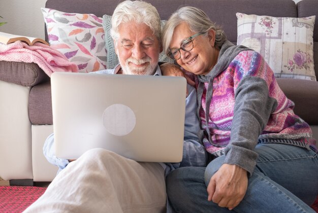 Atrakcyjna para starszych w domu przy użyciu tego samego laptopa razem. Uśmiechnięty starszy emeryt cieszący się wolnym czasem, technologią i mediami społecznościowymi