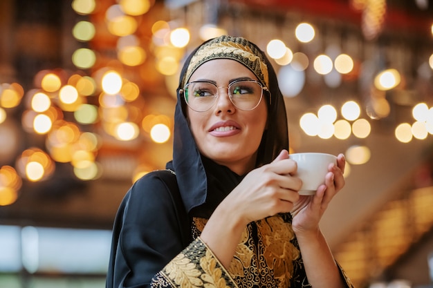 Atrakcyjna pozytywna uśmiechnięta muzułmańska kobieta w tradycyjnym stroju siedzi sama w kawiarni