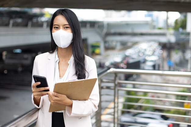 Atrakcyjna profesjonalna azjatycka kobieta biznesu korzystająca z telefonu komórkowego w masce ochronnej zapobiega wirusowi covid-19 w mieście na zewnątrz
