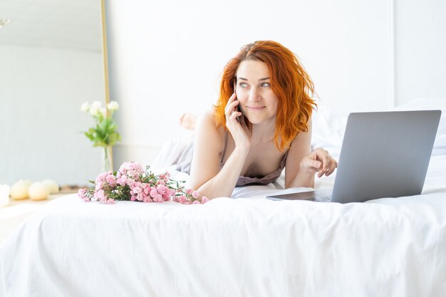 Atrakcyjna ruda kobieta w średnim wieku w piżamie leżąca w łóżku przy użyciu telefonu i laptopa