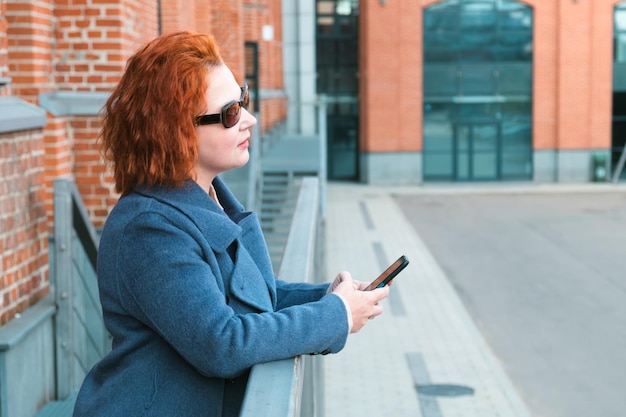 Atrakcyjna rudowłosa kobieta patrzy na ekran smartfona na tle budynku z cegły