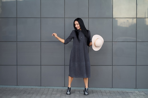 Atrakcyjna śmieszna młoda kobieta jest ubranym młodzieżową szarą obszerną suknię i modne buty trzyma stylowego eleganckiego kapelusz. Model w modnym stroju pozowanie na szarej ścianie miejskiej Portret na zewnątrz.