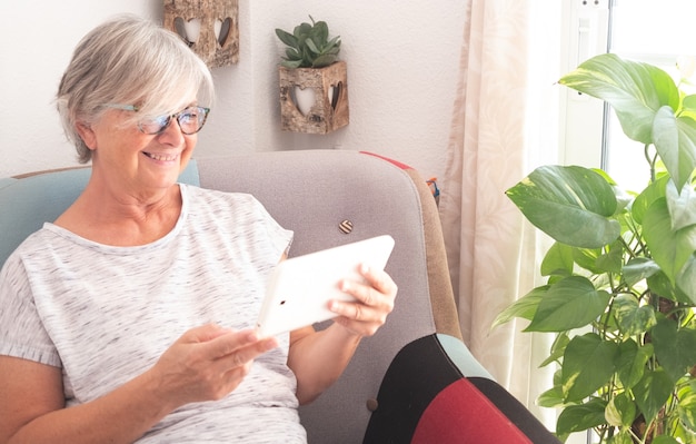 Atrakcyjna starsza kobieta siedzi na fotelu i patrząc na tablet. Siwe włosy, okulary i szeroki uśmiech. Tło z naturalnych roślin i okna. Tylko jedna osoba