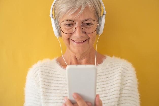Atrakcyjna starsza kobieta w białym swetrze ze słuchawkami podczas korzystania z telefonu, aby obejrzeć film starej generacji i nowej technologii