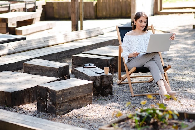 Zdjęcie atrakcyjna, stylowa uśmiechnięta kobieta pracująca, pisząc na laptopie, freelancer siedzący w parku na krześle centrum biznesowe w miejskim stylu ulicznym, edukacja online