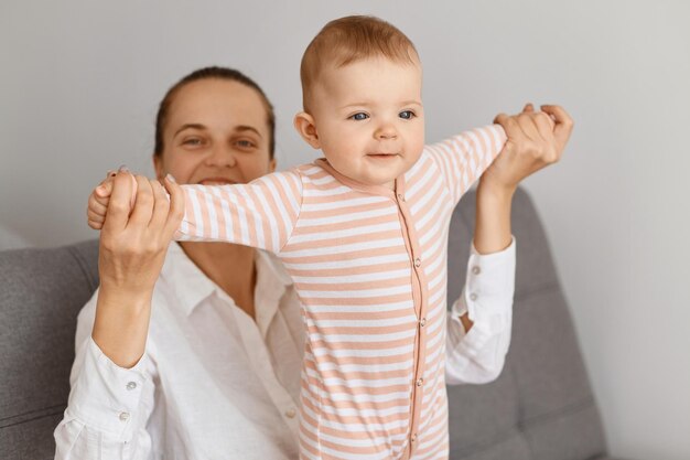 Atrakcyjna Szczęśliwa Mama Bawiąca Się Z Córką W Domu, Trzymająca Za Ręce Dzieciaka I Uśmiechnięta Radośnie, Macierzyństwo I Dzieciństwo, śliczne Urocze Niemowlę Z Mamusią.