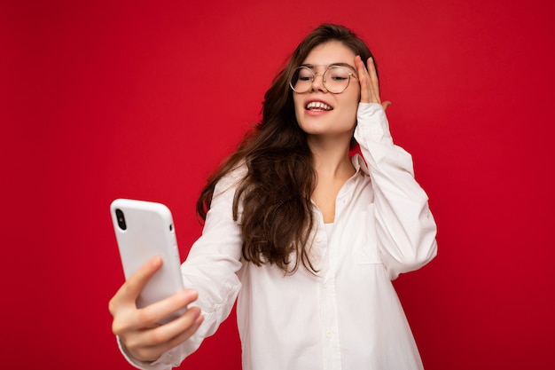 Atrakcyjna, urocza młoda uśmiechnięta szczęśliwa kobieta trzymająca telefon komórkowy i korzystająca z telefonu komórkowego, biorąca selfie w stylowych ubraniach izolowanych na tle ściany