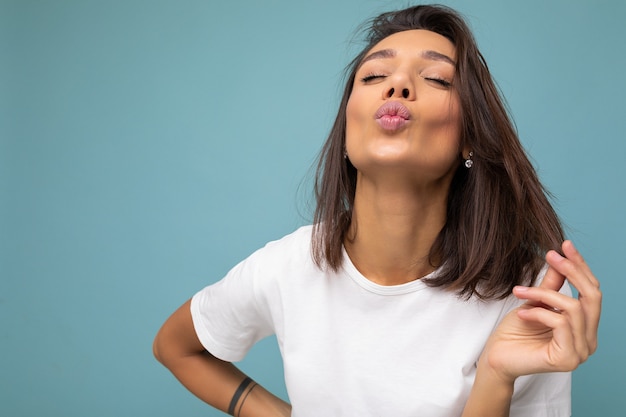 Atrakcyjna Urocza Urocza ładna Seksowna Młoda Brunetka W Przypadkowej Białej Koszulce Do Makiety Na Białym Tle Na Niebieskim Tle Z Wolną Przestrzenią I Dając Buziaka.