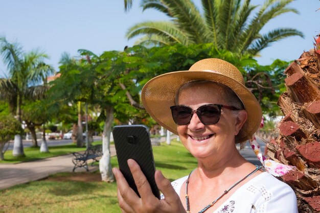 Atrakcyjna uśmiechnięta emerytowana kobieta w publicznym parku za pomocą smartfona, aby porozmawiać z przyjaciółmi, aktywny tryb życia dla emeryta ludzie w przyrodzie pod słońcem