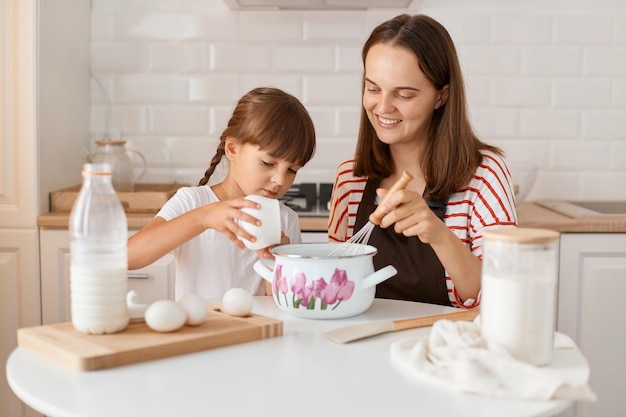 Atrakcyjna zadowolona młoda mama i jej mała córeczka pieką w kuchni w domu, przygotowując domowe ciasto razem, wyrażając szczęście kobieta dziecko ad cukier