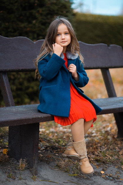 Atrakcyjne dziecko siedzące na ławce w parku w czerwonej sukience i niebieskim płaszczu