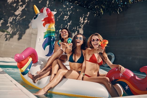 Atrakcyjne młode kobiety w strojach kąpielowych uśmiechają się i celują pistoletami na wodę, unosząc się na nadmuchiwanym jednorożcu w basenie na zewnątrz