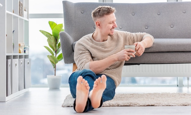 Atrakcyjny blondyn siedzi na dywanie, trzymając w rękach filiżankę herbaty i uśmiechnięty