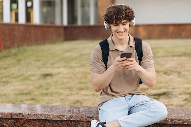 Atrakcyjny kędzierzawy młody mężczyzna na uniwersytecie lub student z telefonem spacerujący po kampusie
